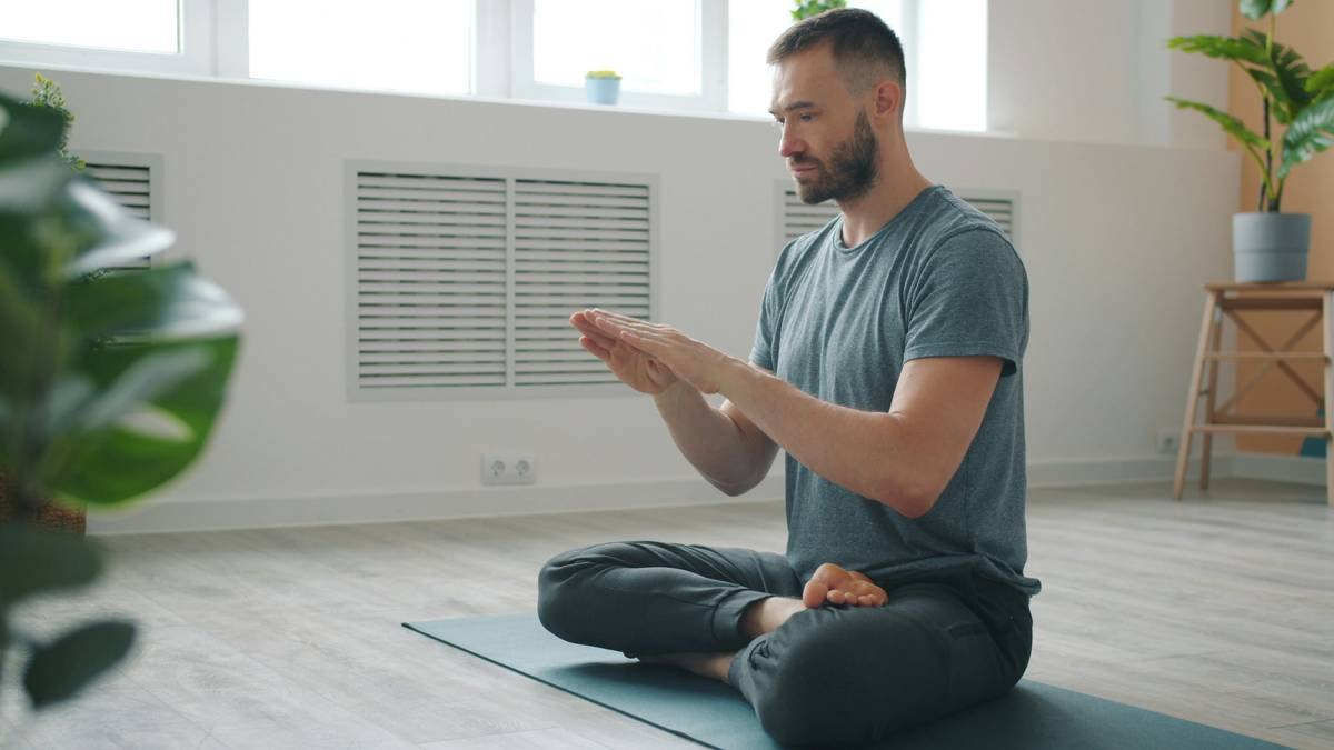 Slow motion of serious young man meditating at home moving hands in namaste with closed eyes sitting on yoga man in cozy apartment. People and relaxation concept.